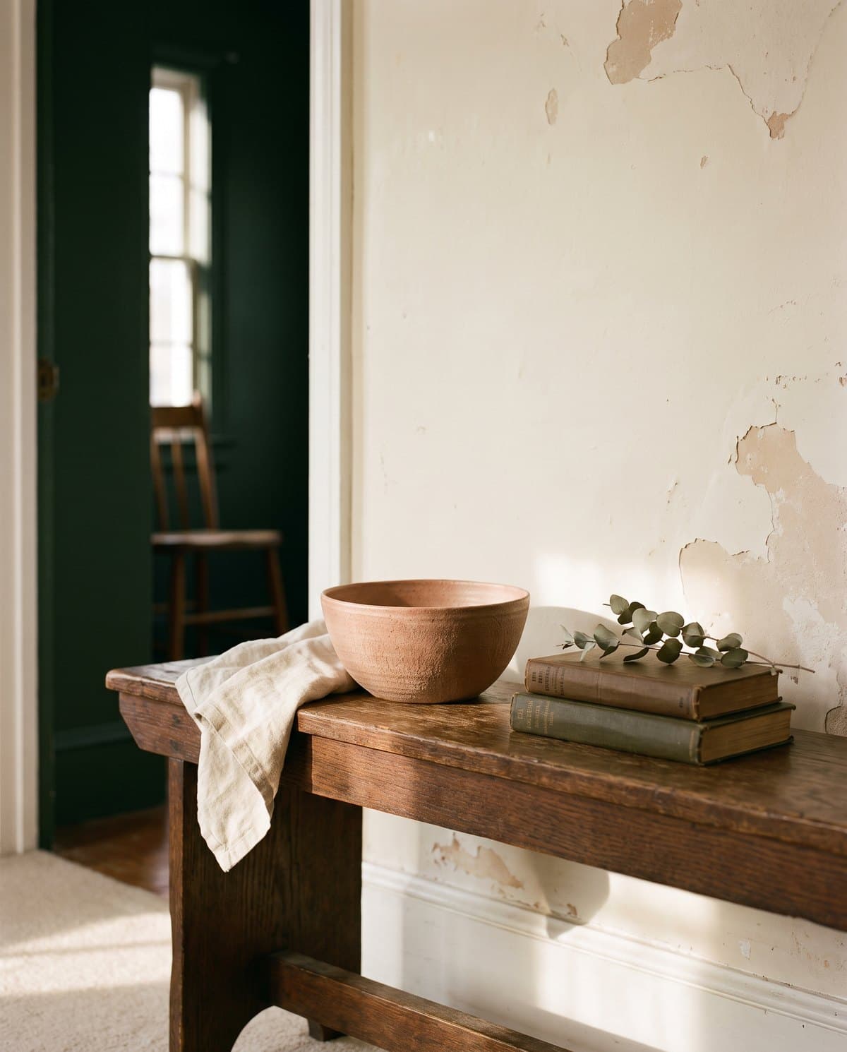 A stoneware bowl, folded linen and a small stack of books on a worn oak bench in late afternoon light.